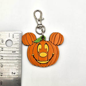 Op de afbeelding: Oranje pompoenvormige sleutelhanger met een lachend gezicht en Mickey Mouse oren. De sleutelhanger is bevestigd aan een zilverkleurige sluiting. De pompoen meet ongeveer 5 cm in hoogte.