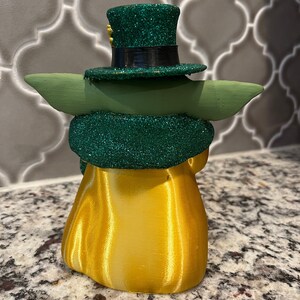 May include: A green and gold figurine of a character wearing a green hat with a gold band and a gold trim. The figurine is wearing a green and gold robe.