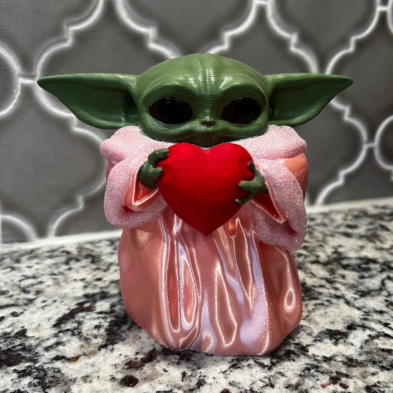 May include: A green and pink figurine of a character holding a red heart. The character has large eyes and a small mouth. The figurine is made of plastic and is about 10 inches tall.