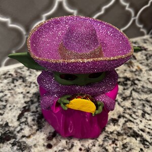 Puede incluir: Una figurita verde y morada de un personaje que lleva un sombrero rosa y sostiene un taco amarillo. La figurita est&aacute; cubierta de purpurina.