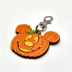 Op de afbeelding: Oranje pompoenvormige sleutelhanger met een lachend gezicht en Mickey Mouse oren. De sleutelhanger heeft een zwarte rand en een zilveren sluiting. De pompoen heeft een groene stengel en gele gezichtskenmerken. Een feestelijk Halloween-accessoire.