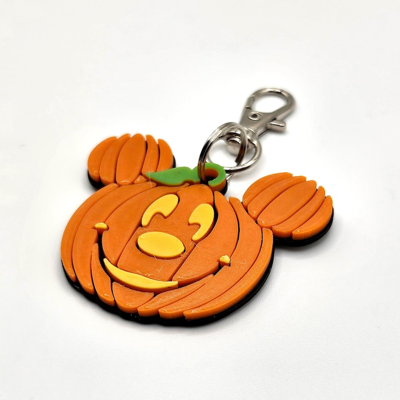 Op de afbeelding: Oranje pompoenvormige sleutelhanger met een lachend gezicht en Mickey Mouse oren. De sleutelhanger heeft een zwarte rand en een zilveren sluiting. De pompoen heeft een groene stengel en gele gezichtskenmerken. Een feestelijk Halloween-accessoire.