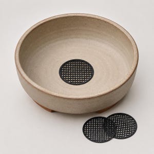 May include: A round, beige ceramic bowl with a speckled texture and a low profile. A circular black plastic mesh grid is placed in the center of the bowl. Two additional identical black circular mesh grids are positioned outside the bowl.