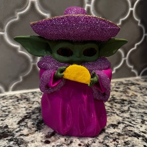 Puede incluir: Una figurita verde y morada de un personaje sosteniendo un taco amarillo. El personaje lleva un sombrero morado con purpurina y una t&uacute;nica morada con purpurina.