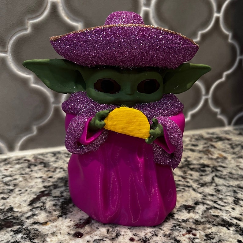 Puede incluir: Una figurita verde y morada de un personaje sosteniendo un taco amarillo. El personaje lleva un sombrero morado con purpurina y una t&uacute;nica morada con purpurina.