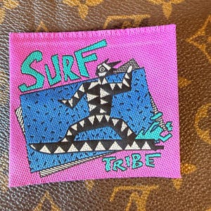 May include: A square embroidered patch featuring a bright pink background with the text SURF in teal at the top and TRIBE in teal at the bottom. A central blue rectangle contains a black and white geometric figure riding a surfboard.