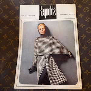 May include: A vintage Reynolds New Collection Designer Coats advertisement featuring a person wearing a black and white houndstooth patterned cape and coat, a dark head covering, and black gloves, set against a plain gray background.