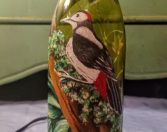 Puede incluir: Una botella de vidrio decorativa con un diseño de pájaro carpintero pintado a mano. La botella presenta una ilustración detallada de un pájaro carpintero posado en una rama de árbol, rodeado de hojas y follaje verdes. La parte superior está envuelta con cuerda.
