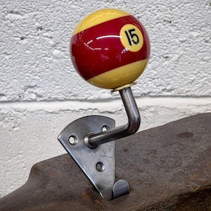 Peut inclure: Un crochet mural unique avec un design de boule de billard. Le crochet présente une boule de billard numéro 15 rouge et jaune, montée sur un support métallique avec un crochet incurvé. Le support est fixé à une base métallique.
