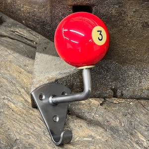 Peut inclure: Un crochet en métal avec une boule de billard rouge portant le numéro 3. Le crochet est fixé à une plaque métallique avec trois trous de vis. Le crochet est monté sur une surface en bois vieilli.
