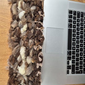 May include: A rectangular, hand-woven textile in shades of brown, cream, and tan, with a textured, looped design. The textile is next to a silver laptop, showing the keyboard.