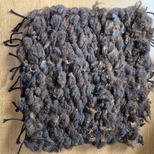 May include: A square, hand-woven textile in shades of brown and gray. The textured surface is made of thick, looped yarn, with black yarn tassels along the edges. The craft item is displayed on a light wood surface.