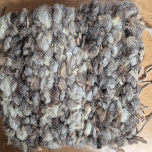 May include: A handwoven, square-shaped textile in shades of gray, brown, and cream. The texture is thick and nubby, with visible loops and knots. Beige cords extend from one corner. This item is suitable for home decor.