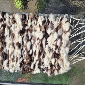 May include: Handwoven textile in a rectangular shape, featuring a textured pattern of cream, brown, and dark brown wool. White threads extend from one edge. The craftwork showcases a rustic, natural aesthetic, suitable for home decor.