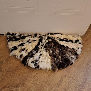May include: A half-circle rug made of thick, fluffy yarn in shades of white, black, and brown. The rug is placed on a wooden floor.