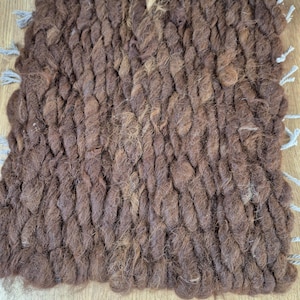 May include: A square, hand-woven brown rug with a textured, rope-like appearance. The rug has a dense weave and is trimmed with gray tassels on the corners. The rug is made of natural fibers and is suitable for indoor use.