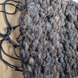 May include: A handmade, textured, square-shaped item with a woven appearance. The item is primarily dark gray with lighter brown and blue-gray accents. Black cords are attached to the item, with tassels at the ends. The item is resting on a light brown surface.