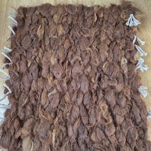 May include: A square, hand-woven rug in shades of brown. The rug has a thick, textured appearance with visible knots and loops. Gray tassels are attached to the corners. This rug is suitable for home decor.