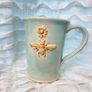 May include: A light blue ceramic mug with a raised bee and flower design. The bee is yellow and the flower is white.
