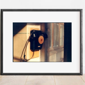 May include: A black wall-mounted telephone with a circular orange dial center, mounted on a light-colored wooden rectangular base. The telephone features a handset resting on top and a black cord hanging down against a light-colored wall.