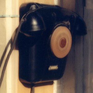 May include: A black wall-mounted telephone with a circular light-colored dial and a handset resting on top. The device features a rectangular base, a black cord extending from the side, and a small rectangular label plate at the bottom.