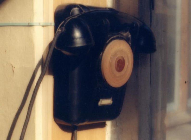 May include: A black wall-mounted telephone with a circular light-colored dial and a handset resting on top. The device features a rectangular base, a black cord extending from the side, and a small rectangular label plate at the bottom.