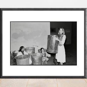 Puede incluir: Fotografía en blanco y negro enmarcada que presenta a varias personas con botes de basura de metal. Una persona sostiene un bote de basura grande, mientras que otras están sentadas cerca de otros más pequeños. La imagen es una toma de estilo documental vintage.