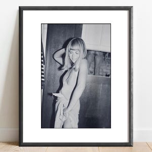 May include: Black and white framed photograph. The photograph features a person in a light-colored dress, leaning against a wooden door. The person is smiling and has their hair styled in a bob.