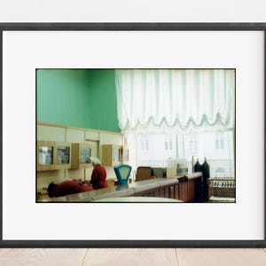 May include: A framed photograph depicting an interior scene with mint green walls, light-colored wooden cabinets, and white draped curtains. Two figures in red clothing are positioned at a counter with a blue scale and dark canisters.