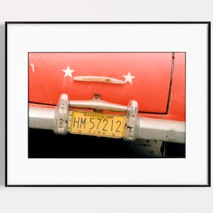 May include: Close-up of a vintage red car's rear. Features a chrome bumper, a yellow license plate with the text "HM 57212 CUBA", and two white star decorations. The car's paint shows signs of age.