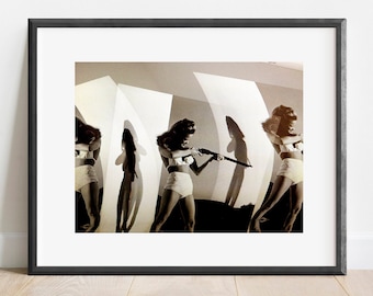 May include: A framed black and white photographic print featuring a repeating sequence of a figure in a two-piece swimsuit holding a long rifle. The composition includes curved white panels and dramatic shadows against a light background.