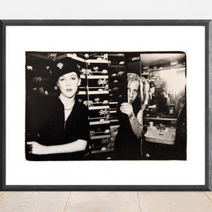 May include: Black and white framed photograph featuring two people in a bathroom setting. One person wears a hat and dark clothing, while the other is partially visible, holding a patterned divider. The background includes patterned wallpaper and a mirror.