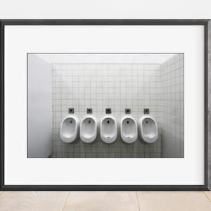 May include: A framed wall art print featuring a row of five white ceramic urinals mounted on a wall covered in white square tiles. Each urinal is equipped with a small, dark, square-shaped flush mechanism positioned directly above it.