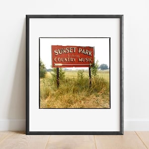 May include: Framed print of a vintage sign for Sunset Park featuring Country Music. The sign is red with white lettering and an arrow, set against a field and sky. The print is in a black frame.