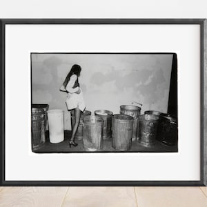 May include: A black and white photograph of a woman standing in front of a wall with a row of dustbins in front of her.