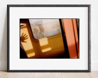 May include: A framed photographic print featuring a view from inside a train carriage. The image captures a golden-hued interior wall with a rectangular window showing a blurred landscape, and the distinct shadow of a hand cast upon the yellow surface.