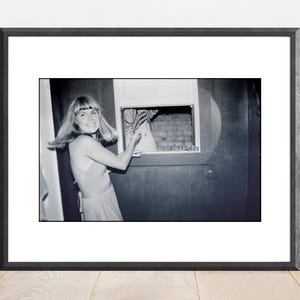 May include: A black and white photo of a woman in a dress standing in front of a window. She is looking to the right and smiling. The window is open and there is a view of a building in the background.