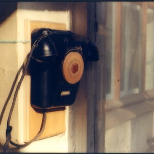 May include: A black wall-mounted telephone with a circular rotary dial featuring a light wood-toned center. The device is attached to a rectangular light-colored wooden mounting board and includes a black handset resting on top.