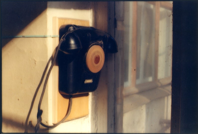 May include: A black wall-mounted telephone with a circular rotary dial featuring a light wood-toned center. The device is attached to a rectangular light-colored wooden mounting board and includes a black handset resting on top.