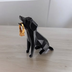 May include: A black, geometric dog figurine holding a gold ring in its mouth. The dog is sitting on a light wood surface.