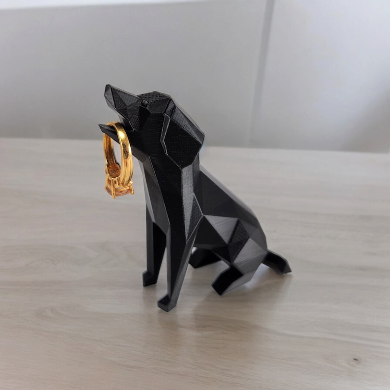 May include: A black, geometric dog figurine holding a gold ring in its mouth. The dog is sitting on a light wood surface.