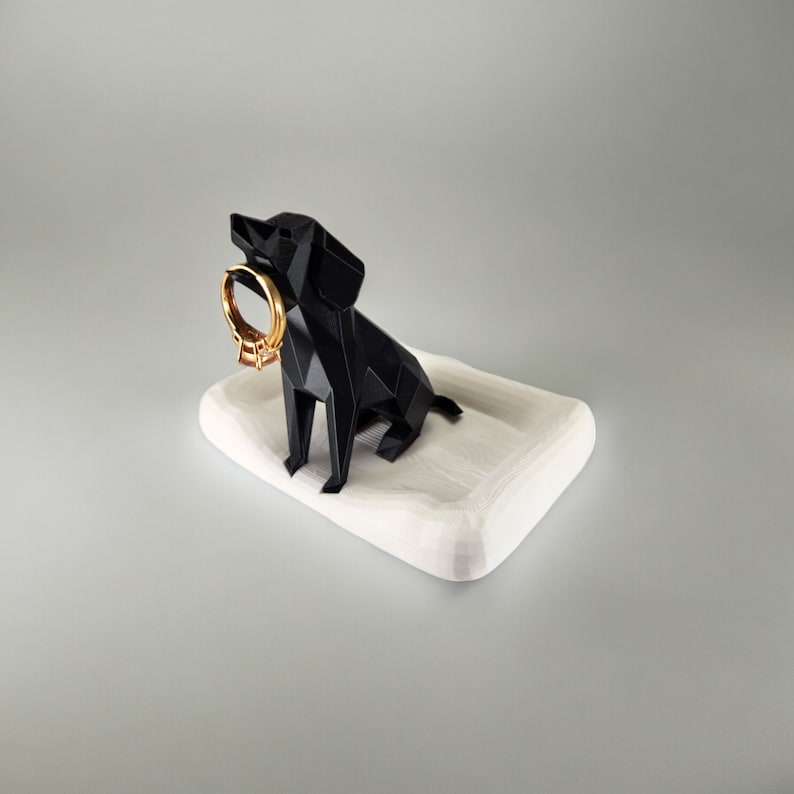 May include: A black geometric dog figurine holding a gold ring, sitting on a white ceramic tray. The dog is a ring holder.