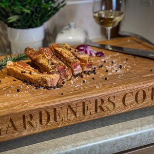 Puede incluir: Una tabla de cortar de madera grande con las palabras "Gardeners Cottage" grabadas en la parte delantera. La tabla est&aacute; cubierta de sal y pimienta y tiene varios trozos de carne cocida encima.