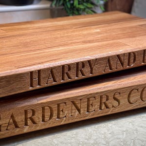 Puede incluir: Dos tablas de cortar de madera apiladas una encima de la otra. La tabla superior est&aacute; grabada con "HARRY AND EMMA" y la tabla inferior est&aacute; grabada con "GARDENERS COTTAGE".
