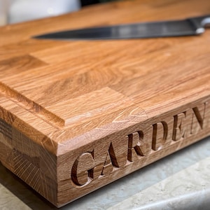 Puede incluir: Una tabla de cortar de madera grande, rectangular, con un borde biselado y una ranura alrededor del per&iacute;metro. La tabla es de madera marr&oacute;n claro y tiene las palabras "GARDENERS CO." grabadas en el borde frontal.