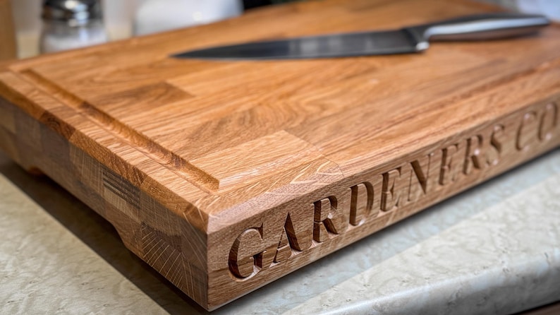 Puede incluir: Una tabla de cortar de madera grande, rectangular, con un borde biselado y una ranura alrededor del per&iacute;metro. La tabla es de madera marr&oacute;n claro y tiene las palabras "GARDENERS CO." grabadas en el borde frontal.