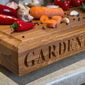 Puede incluir: Una tabla de cortar de madera con el texto "GARDENERS CO." grabado en la parte delantera. La tabla es marr&oacute;n y tiene una veta de madera natural. Hay varias verduras y especias en la tabla, incluyendo pimientos rojos, zanahorias y granos de pimienta.