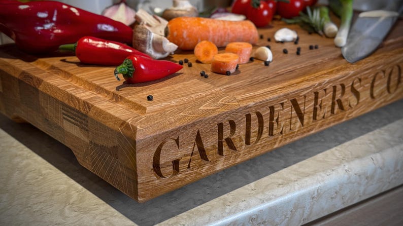 Puede incluir: Una tabla de cortar de madera con el texto "GARDENERS CO." grabado en la parte delantera. La tabla es marr&oacute;n y tiene una veta de madera natural. Hay varias verduras y especias en la tabla, incluyendo pimientos rojos, zanahorias y granos de pimienta.