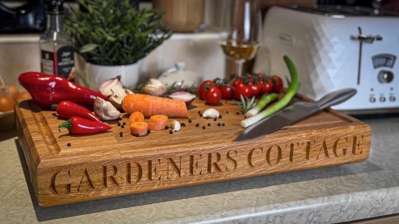 Puede incluir: Una tabla de cortar de madera con las palabras "GARDENERS COTTAGE" grabadas en la parte delantera. La tabla est&aacute; cubierta con varias verduras, incluyendo pimientos rojos, cebollas verdes, zanahorias y tomates.