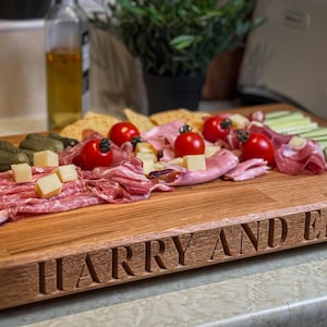 Puede incluir: Una tabla de servir de madera con los nombres "Harry y Emma" grabados en la parte delantera. La tabla est&aacute; llena de una variedad de alimentos, incluyendo salami, queso, uvas rojas y pepinos.